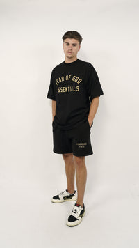 Man wearing Fear of God Essentials FW24 Black Yellow Summer Set with black oversized shirt and shorts paired with black and white sneakers, minimalist studio background.