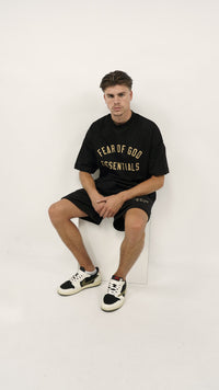 A young man wearing the Fear of God Essentials FW24 Black Yellow Summer Set with black shorts and a matching oversized black t-shirt featuring yellow lettering, paired with stylish black and white sneakers.