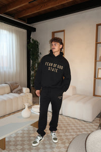 Man wearing a black hoodie and pants with "Fear of God Essentials FW24 Black Yellow State Lange Broek" text, styled with sneakers in a modern living room, The real deal.