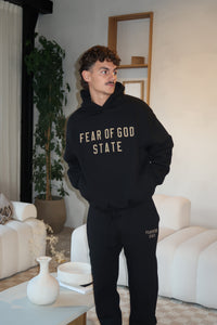 A man wearing the Fear of God Essentials FW24 Black Yellow State Hoodie in a modern living room setting, blending casual style with a cozy atmosphere.