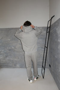 Person wearing the Fear of God Essentials 1977 Dark Oatmeal Full set, shown from the back in a minimalist setting with gray walls and casual sneakers, embodying The real deal.