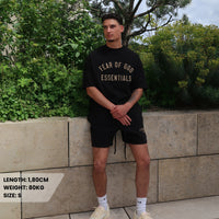 Man wearing Fear of God Essentials FW24 Black Yellow Summer Set with black t-shirt and shorts posing outdoors in casual sneakers, surrounded by greenery.