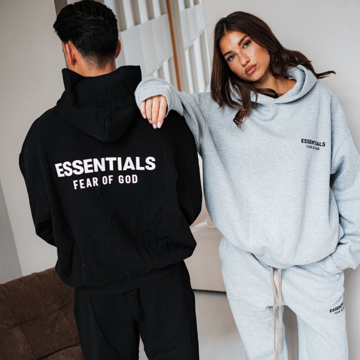 Fear of God Essentials FW24 Light Heather Hoodie, paar samen in grijze en zwarte hoodies met witte ESSENTIALS-print op rug