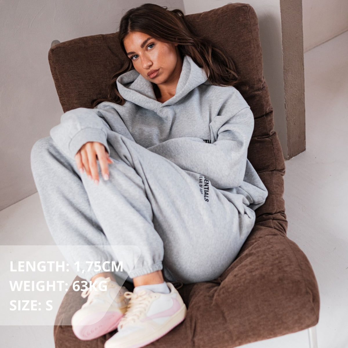 Fear of God Essentials FW24 Light Heather Hoodie, gedragen zittend met grijze hoodie en witte sneakers met roze accenten