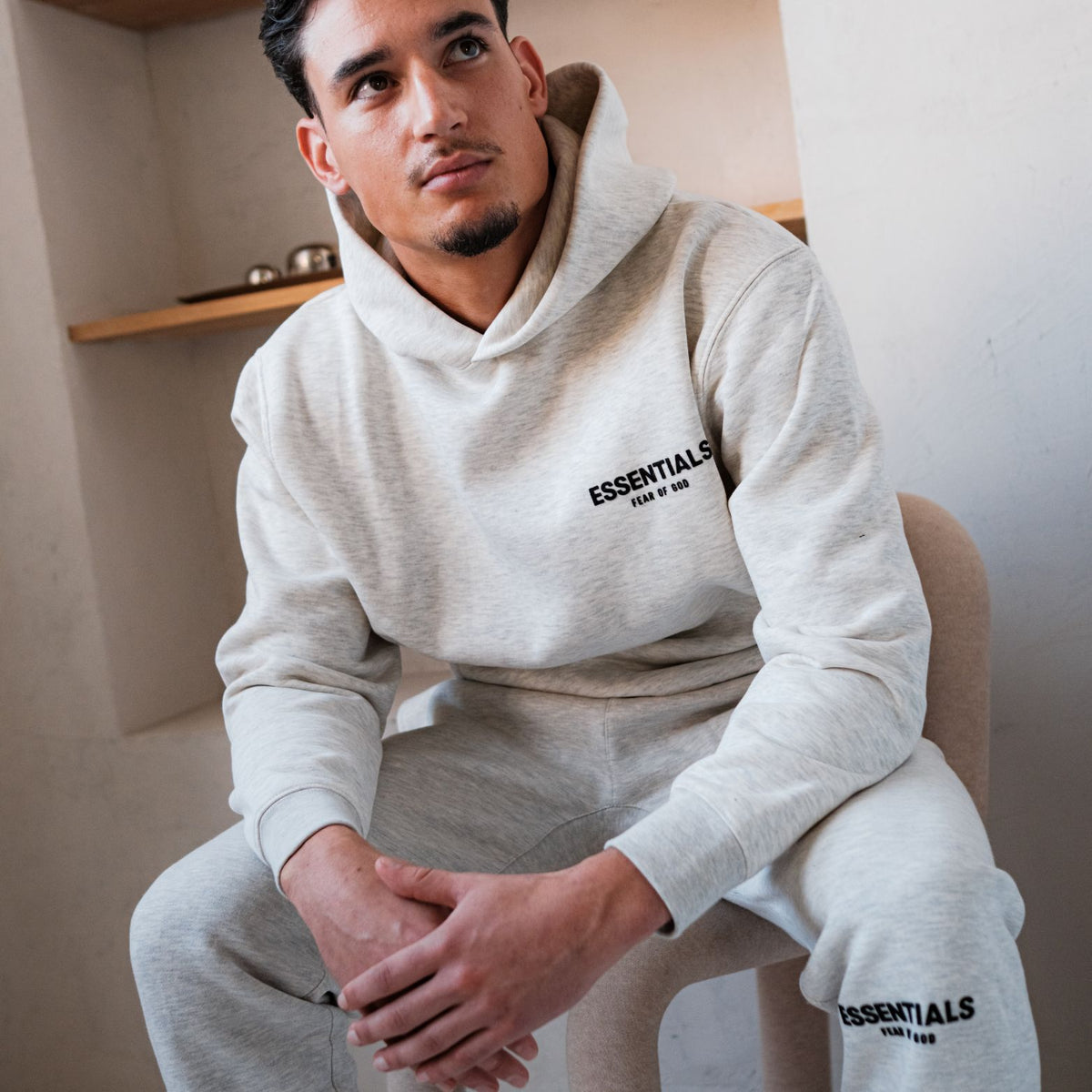 Fear of God Essentials Light Oatmeal Hoodie, vooraanzicht met borststiksel en grijze joggingbroek zichtbaar